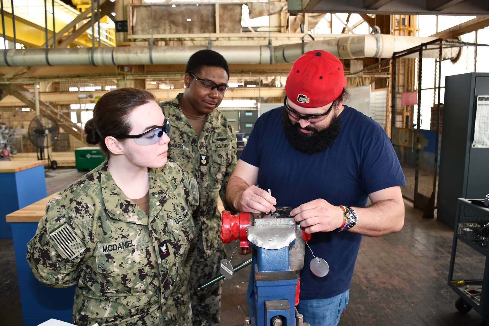 During their annual Navy Reserve two-week-long training period at Norfolk Naval Shipyard (NNSY), SurgeMain Charlotte Machinist Mate Third Class Ashton McDaniel (left) and SurgeMain Atlanta Machinist Mate First Class Kyle Leonard (center) watches NNSY’s Outside Machine Shop (Shop 38) Outside Machinist James Derrickson (right) as he demonstrates how to use a top-loaded ball valve, breaking down, taking measurements, using the center line tool to get the stem center line measurement and giving them a little refresher on some of the precision measuring tools that they would use and the proper tools to take apart the valve.