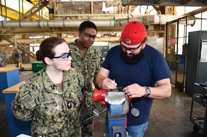 During their annual Navy Reserve two-week-long training period at Norfolk Naval Shipyard (NNSY), SurgeMain Charlotte Machinist Mate Third Class Ashton McDaniel (left) and SurgeMain Atlanta Machinist Mate First Class Kyle Leonard (center) watches NNSY’s Outside Machine Shop (Shop 38) Outside Machinist James Derrickson (right) as he demonstrates how to use a top-loaded ball valve, breaking down, taking measurements, using the center line tool to get the stem center line measurement and giving them a little refresher on some of the precision measuring tools that they would use and the proper tools to take apart the valve.