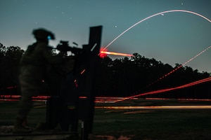 A soldier fires a weapon to create streaks of light against a wooded area and night sky.
