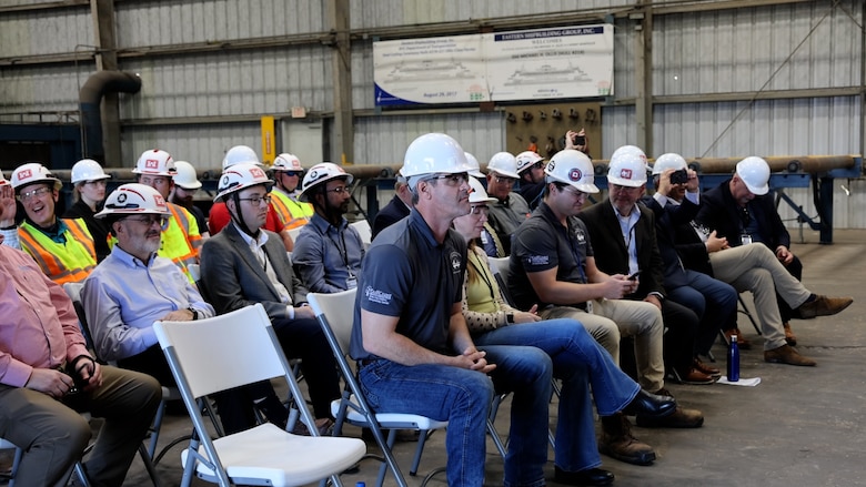 Team members, seated in chairs, listen to remarks during a steel cutting ceremony at a shipyard.