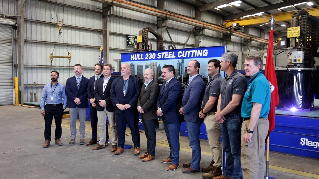 Leaders gather in front of a banner during a steel cutting ceremony at a shipyard.