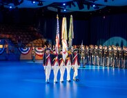 Army Soldiers wearing Revolutionary War-era uniforms are presenting the colors during a ceremony in a large hall. There are other Soldiers wearing modern service dress uniforms who are standing in formation in the background.