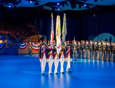 Army Soldiers wearing Revolutionary War-era uniforms are presenting the colors during a ceremony in a large hall. There are other Soldiers wearing modern service dress uniforms who are standing in formation in the background.