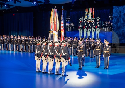 Army Soldiers in various uniforms are standing in formations in a large hall during a ceremony.