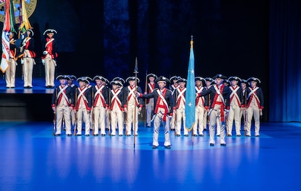 Army Soldiers dressed in Revolutionary War-era uniforms with blue jackets, white pants and black tri-cornered hats, are standing in formation during a ceremony.