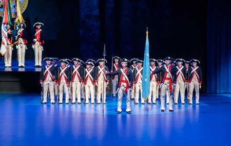 Army Soldiers dressed in Revolutionary War-era uniforms with blue jackets, white pants and black tri-cornered hats, are standing in formation during a ceremony.