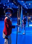 An Army Soldier wearing a red Revolutionary War-era uniform with large furry hat is holding a long staff in his right hand while waiting during a ceremony in a large hall.