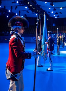 An Army Soldier wearing a red Revolutionary War-era uniform with large furry hat is holding a long staff in his right hand while waiting during a ceremony in a large hall.