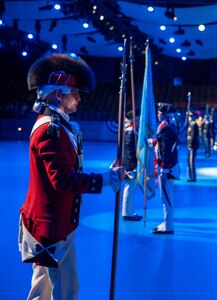 An Army Soldier wearing a red Revolutionary War-era uniform with large furry hat is holding a long staff in his right hand while waiting during a ceremony in a large hall.