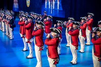 Army Soldiers dressed in red Revolutionary War-era uniforms are standing in rows preforming on musical instruments during a ceremony.