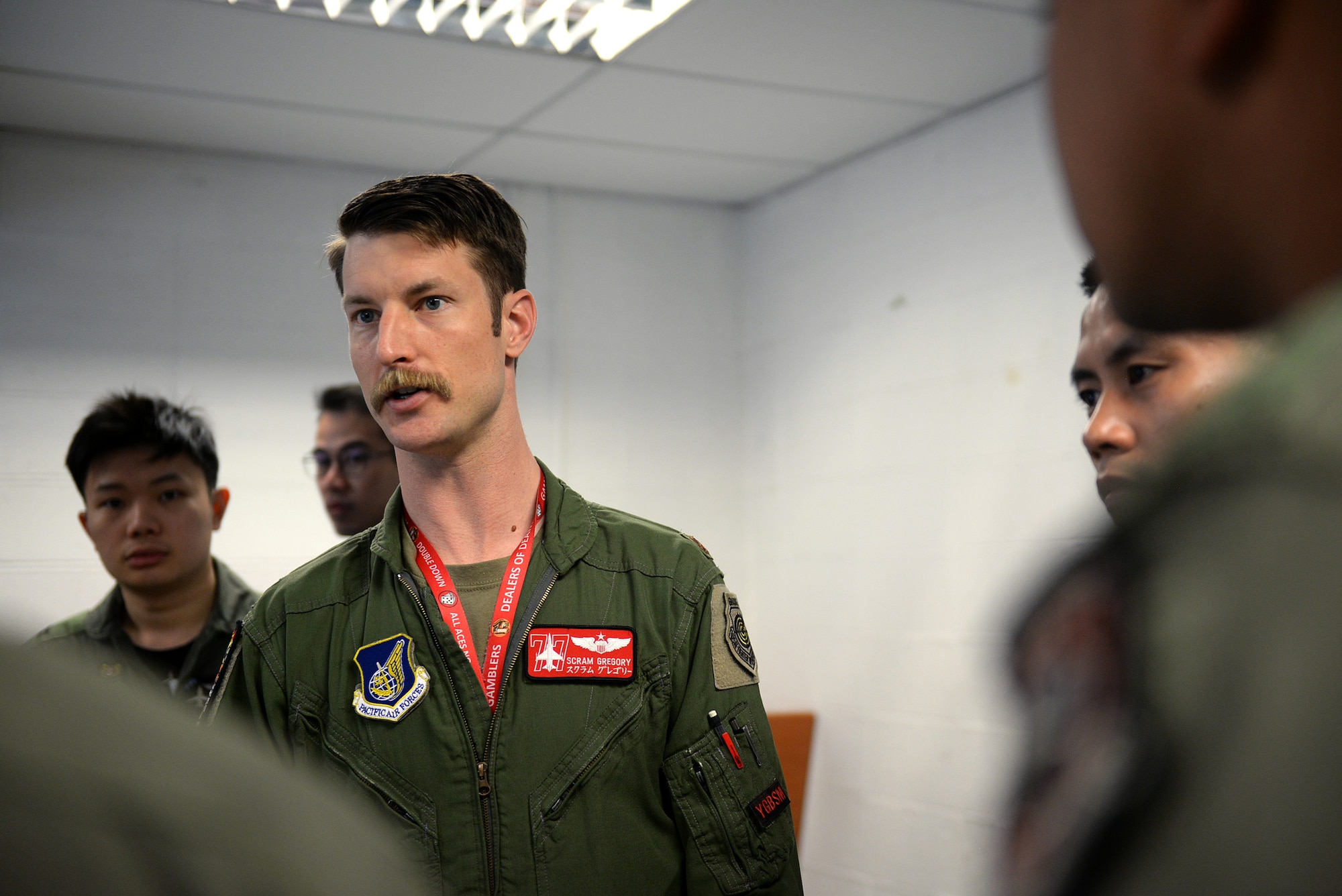 A mustachioed pilot in a jump suit stand in the middle of a room surrounded by similarly dressed pilots