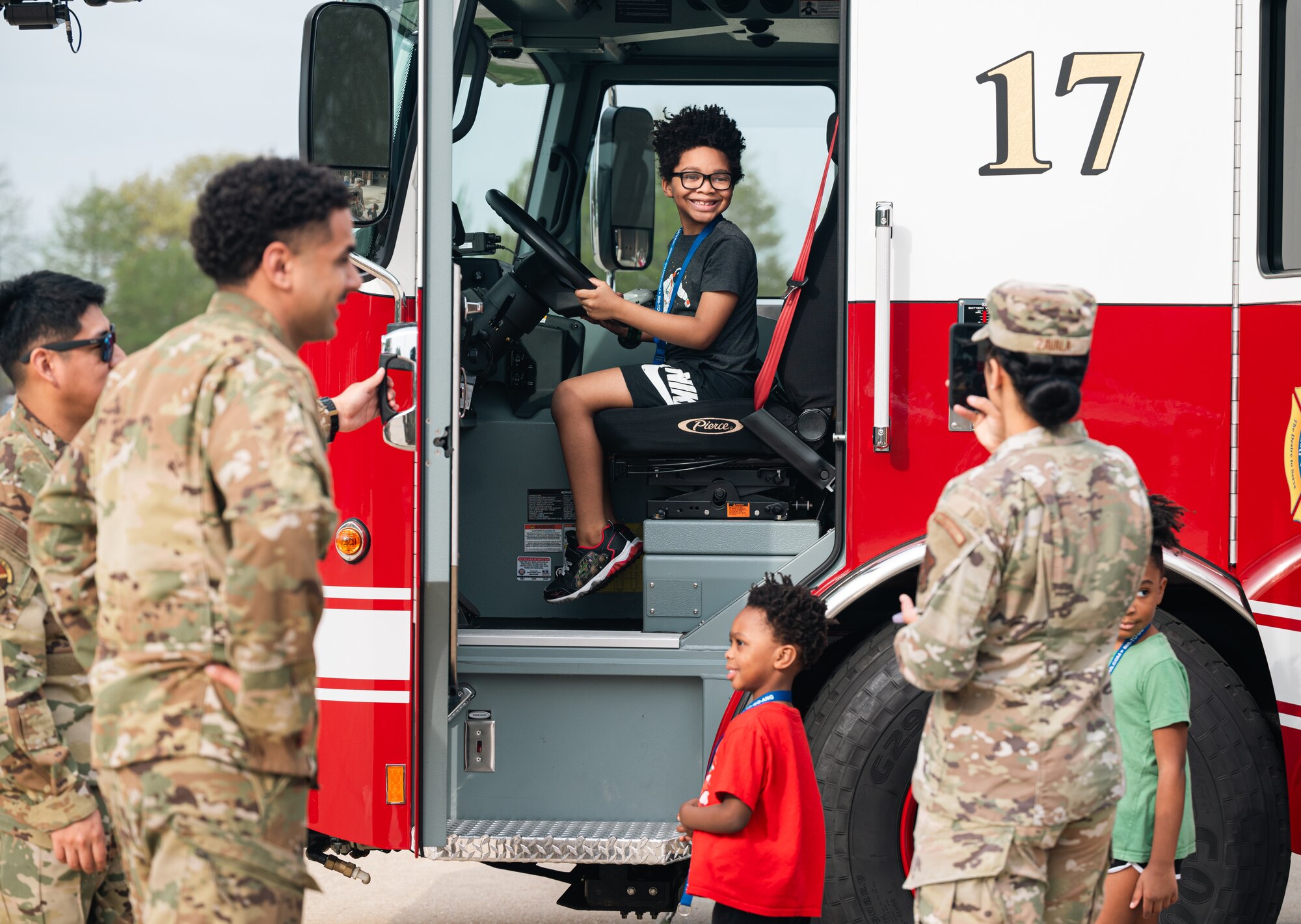 A kid smiles in a firetruck while people stand around outside