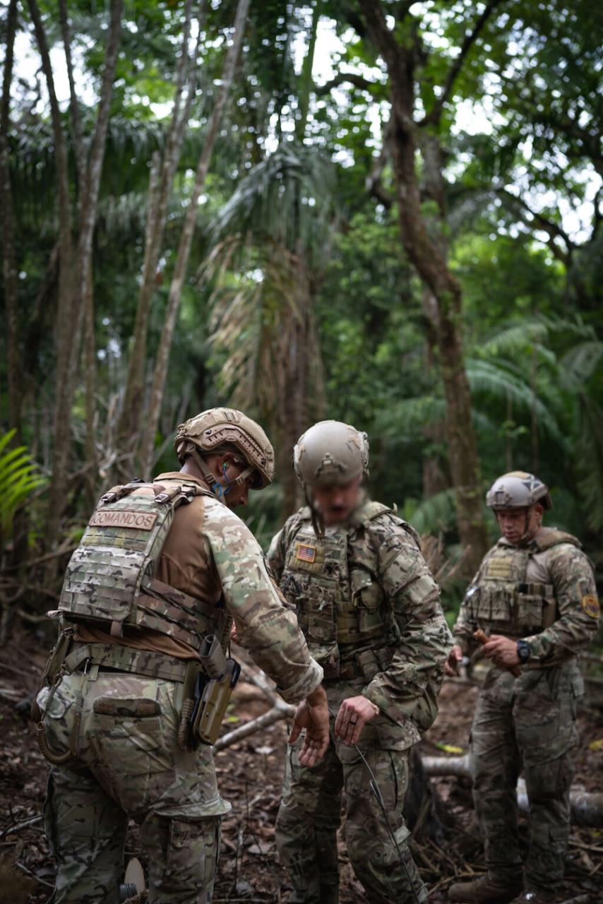 Three men wearing military uniforms stand under trees while working together.