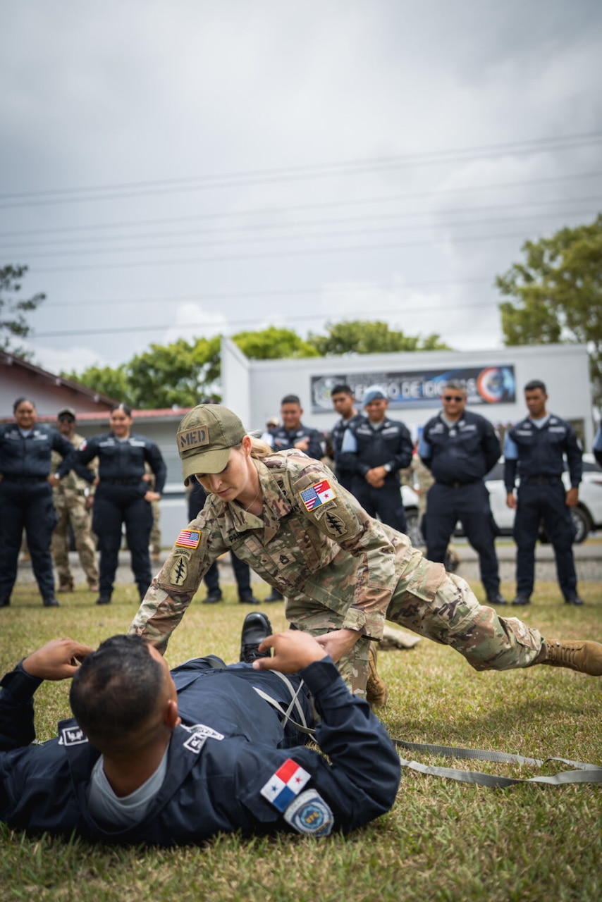A woman wearing an American military uniform kneels over a man wearing a dark blue uniform while he is on the ground. A group of people wearing the same dark blue uniform watch.