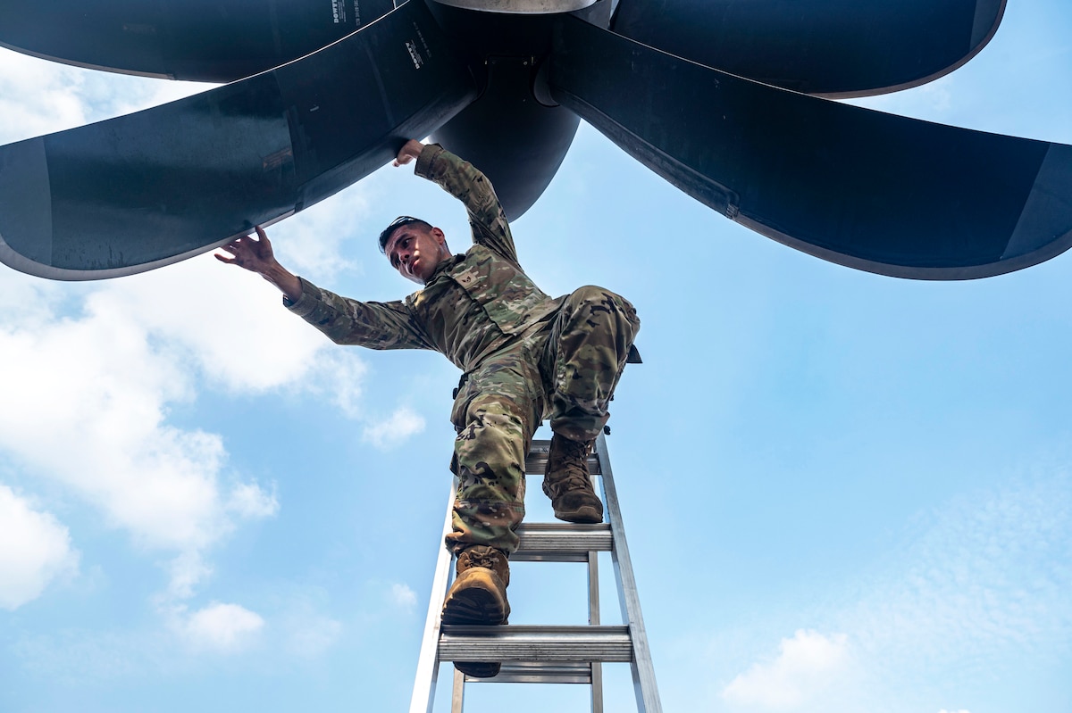 An airman stands at the top of a ladder and reaches up with both arms to examine a blade on an aircraft propeller against a partly cloudy sky.