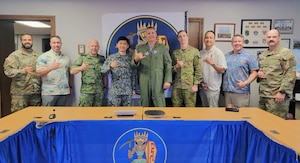 a mix of 9 soldiers and civilians med pose for a photo standing behind a conference table.