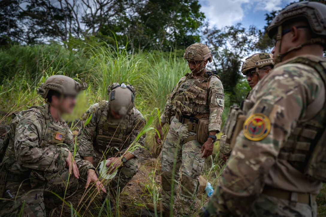 Five men wearing military uniforms are in a field of long grass. One is kneeling while talking the other are in a group around him.