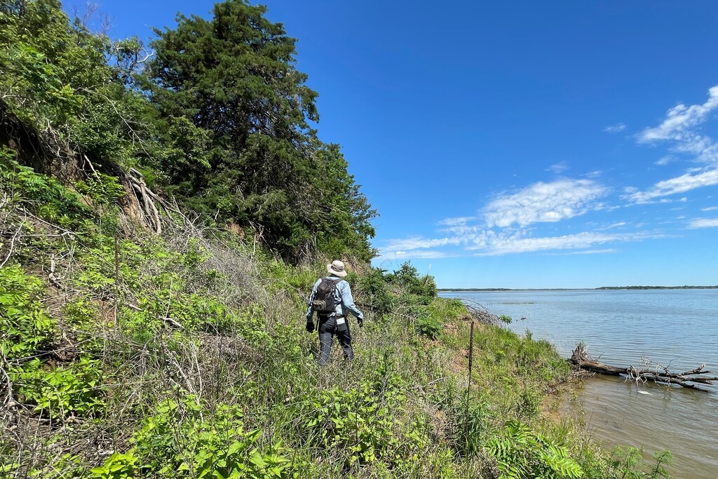 Man wearing backpack and sun hat walking along overgrown lake shoreline