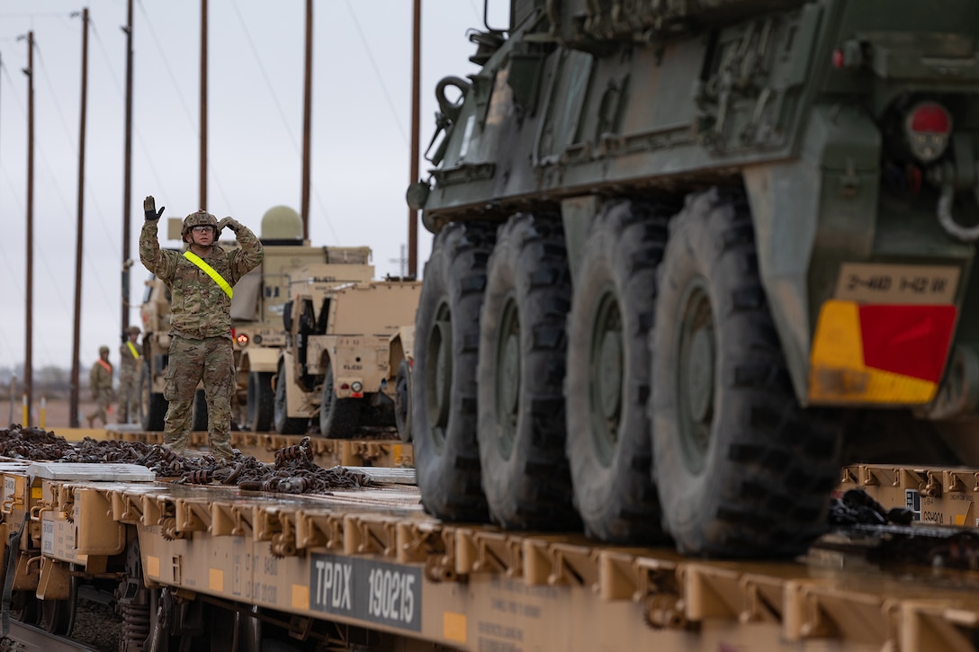 A soldier guides a military vehicle on tracks during an overcast day.