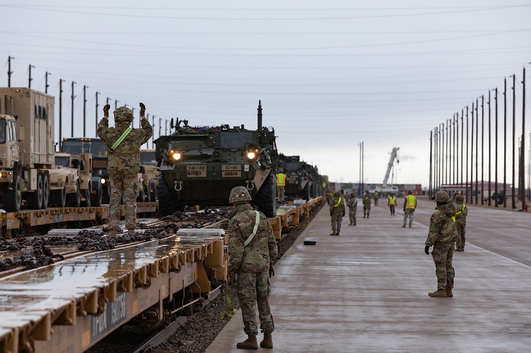 A soldier signals to a line of military vehicles parked on a platform on a rail.