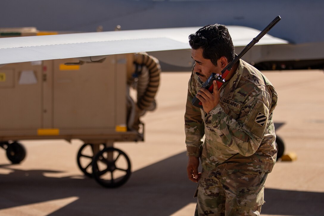 A soldier inspects the wing of a a large drone while holding a radio to his ear.