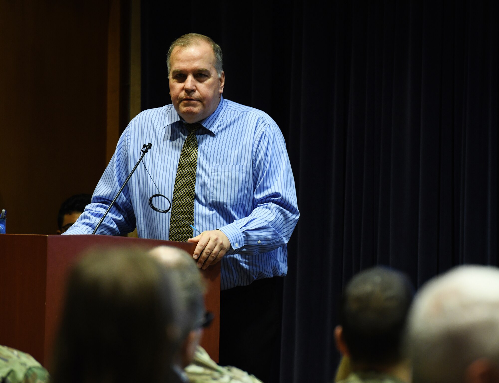 man in dress shirt and tie talks to an audience while standing at a podium.