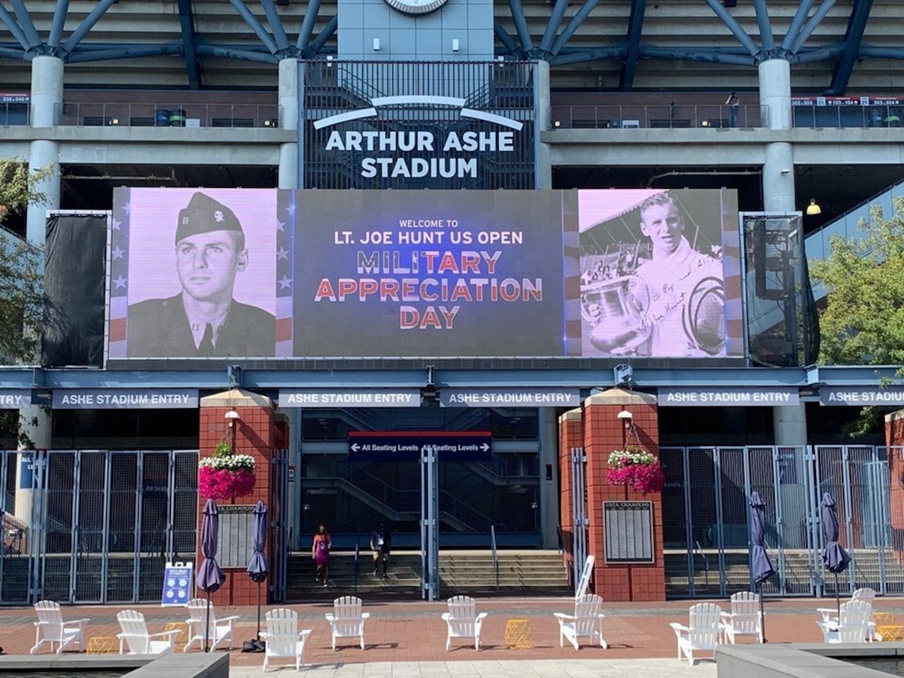 A large electronic billboard displays pictures of a man in a military uniform on the left and a man in athletic attire holding a trophy on the right. The sign reads “Welcome to Lt. Joe Hunt U.S. Open Military Appreciation Day.” An entrance to a stadium is shown.
