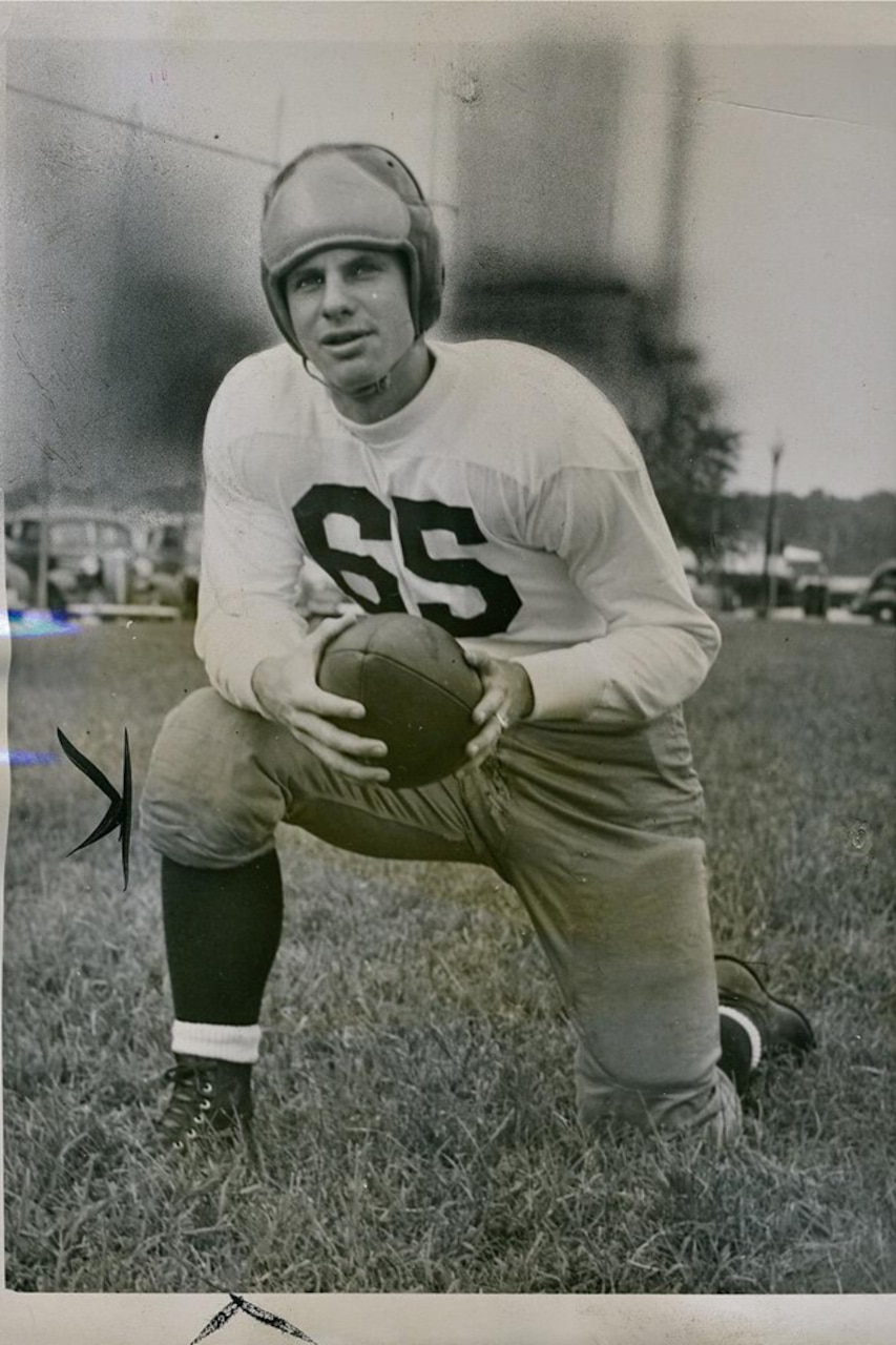 A man in a football uniform takes a knee while holding a football.