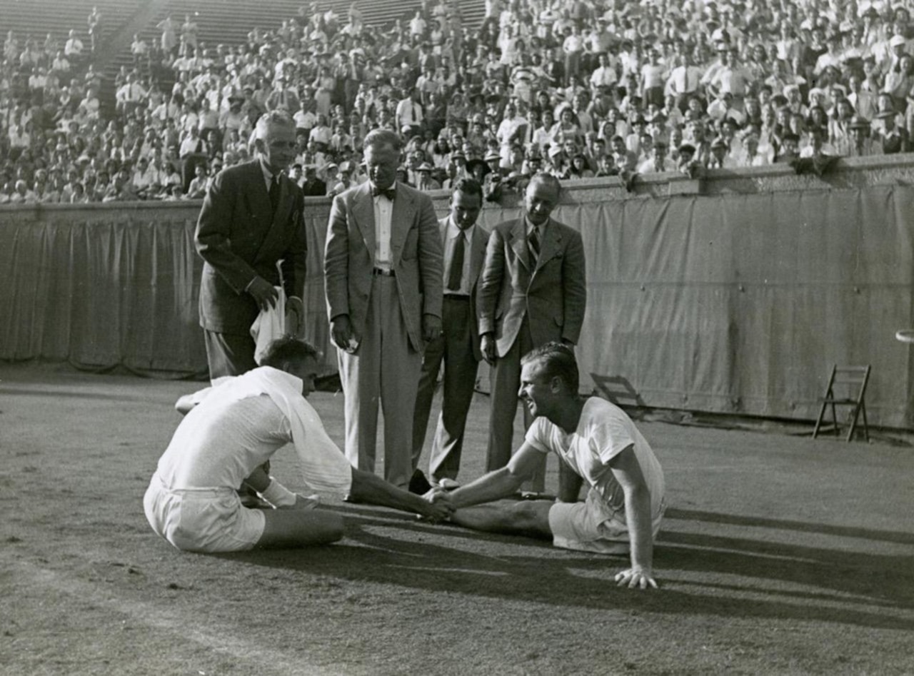 Men in athletic attire shake hands while seated on the ground. Several men in business attire stand behind them, and a large crowd is in the background.