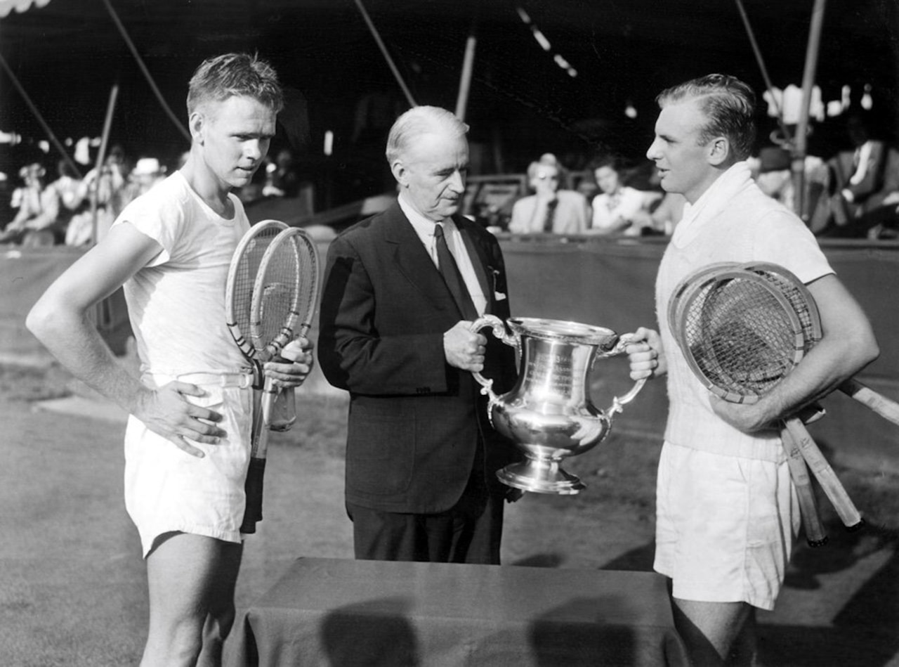 A man in business attire hands a man wearing athletic attire and holding tennis rackets a trophy. Another man wearing athletic attire and holding tennis rackets looks on.