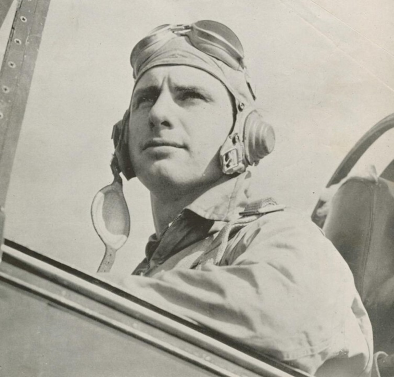 A man in a military uniform is shown in an aircraft cockpit with goggles on his head.