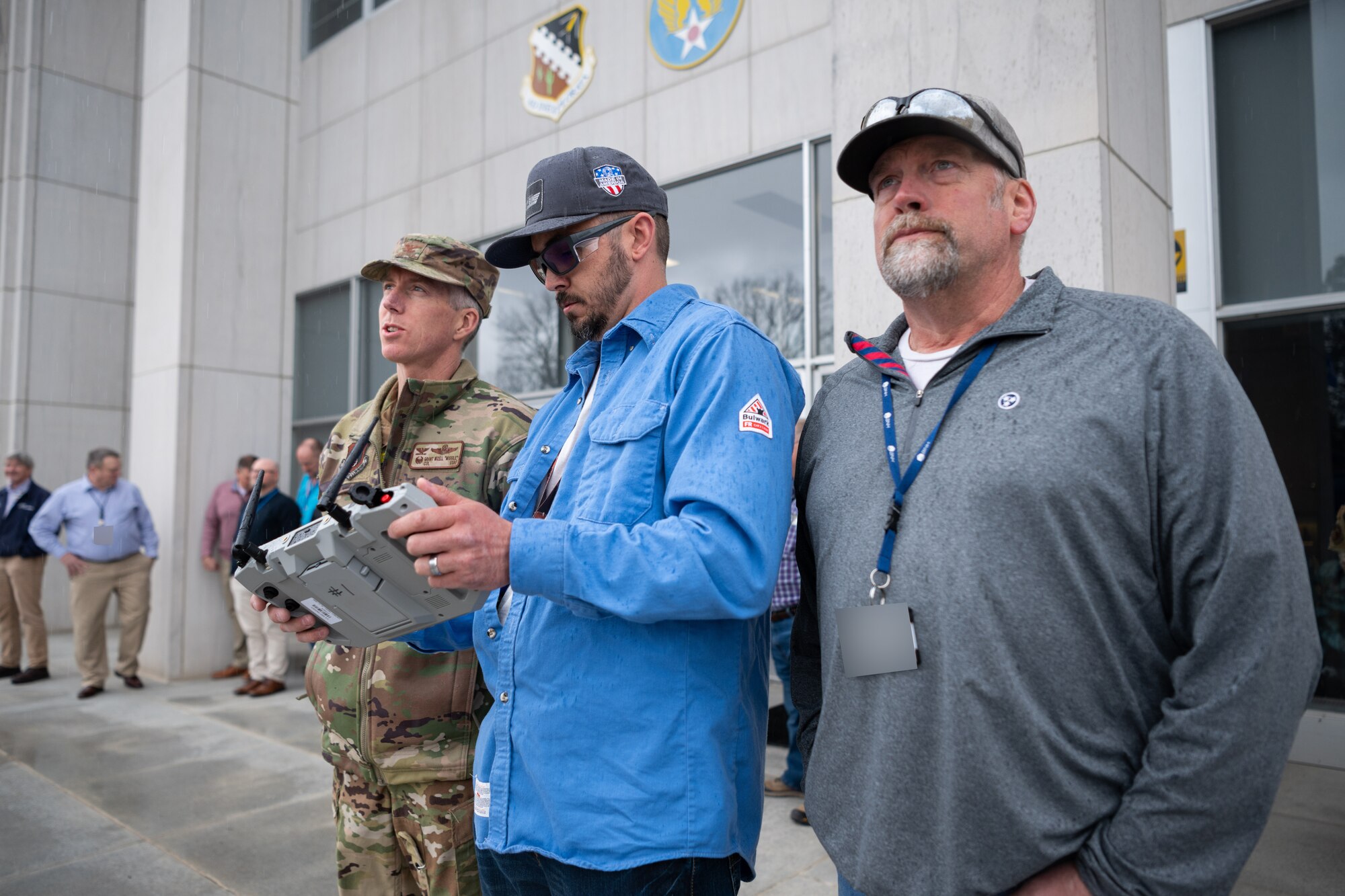 Arnold Engineering Development Complex Commander Col. Grant Mizell, left, watches on while Robert Hastings, sUAS operator, center, and Matt Holder, sUAS observer, pilot a Small Unmanned Aircraft System, also known as an sUAS or a “drone,” above the Administration and Engineering Building at Arnold Air Force Base, Tenn., during a March 5, 2025 demonstration of the device. AEDC is headquartered at Arnold AFB. The sUAS will soon be used at Arnold to perform asset health assessments. The device is equipped with both digital and infrared imaging capabilities and will be flown around installation buildings, plant facilities and associated equipment to ensure there are no compromises to such assets and to assess the structural integrity of facilities. (U.S. Air Force photo by Keith Thornburgh) (This image has been altered by obscuring badges for security purposes)