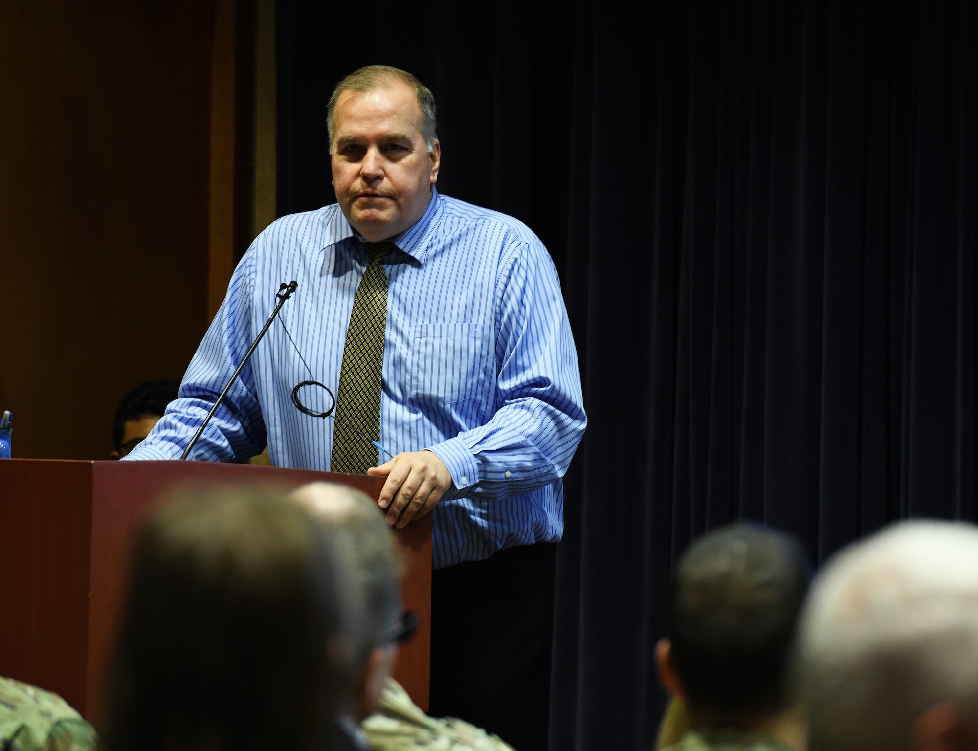 man in dress shirt and tie talks to an audience while standing at a podium.