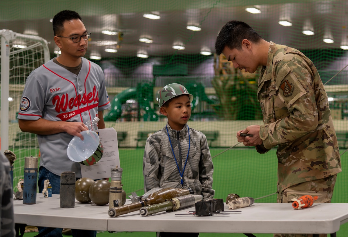 A U.S. Air Force Airman assigned to the 35th Civil Engineer Squadron explosive ordnance disposal flight showcases simulated ordnances to a family during Operation CODE (Children on Deployment Exercise) at Misawa Air Base, Japan, April 5, 2025. Operation CODE bridges the gap between service and family, turning understanding into strength and connection into resilience. (U.S. Air Force photo by Senior Airman Brittany Russell)