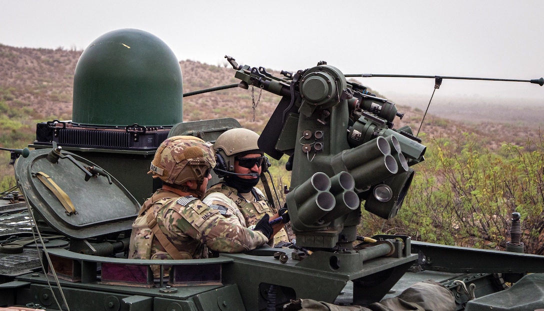 Two soldiers ride on top of a military vehicle.