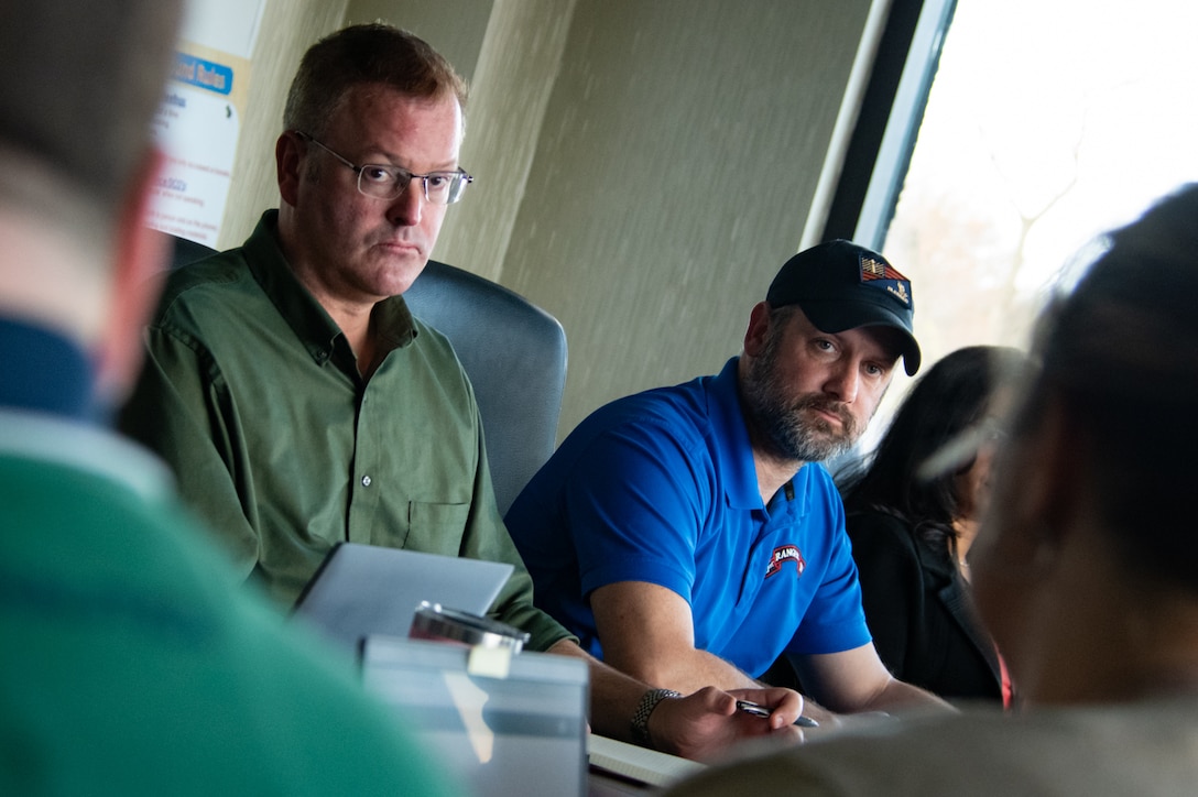 Two men seated at a conference room table look across the table toward others.