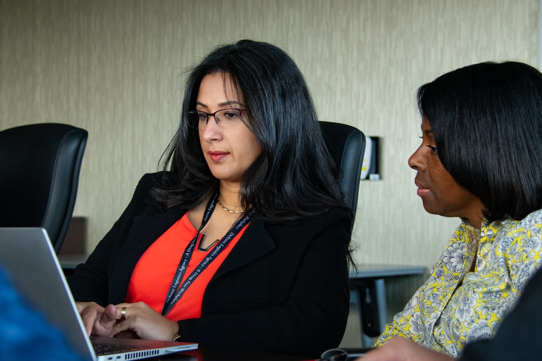 Two women sit at a conference room table looking at a laptop computer.