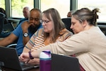 Two women and a man gather around a laptop computer while seated at a conference room table. The woman on the far right is pointing at the computer screen.