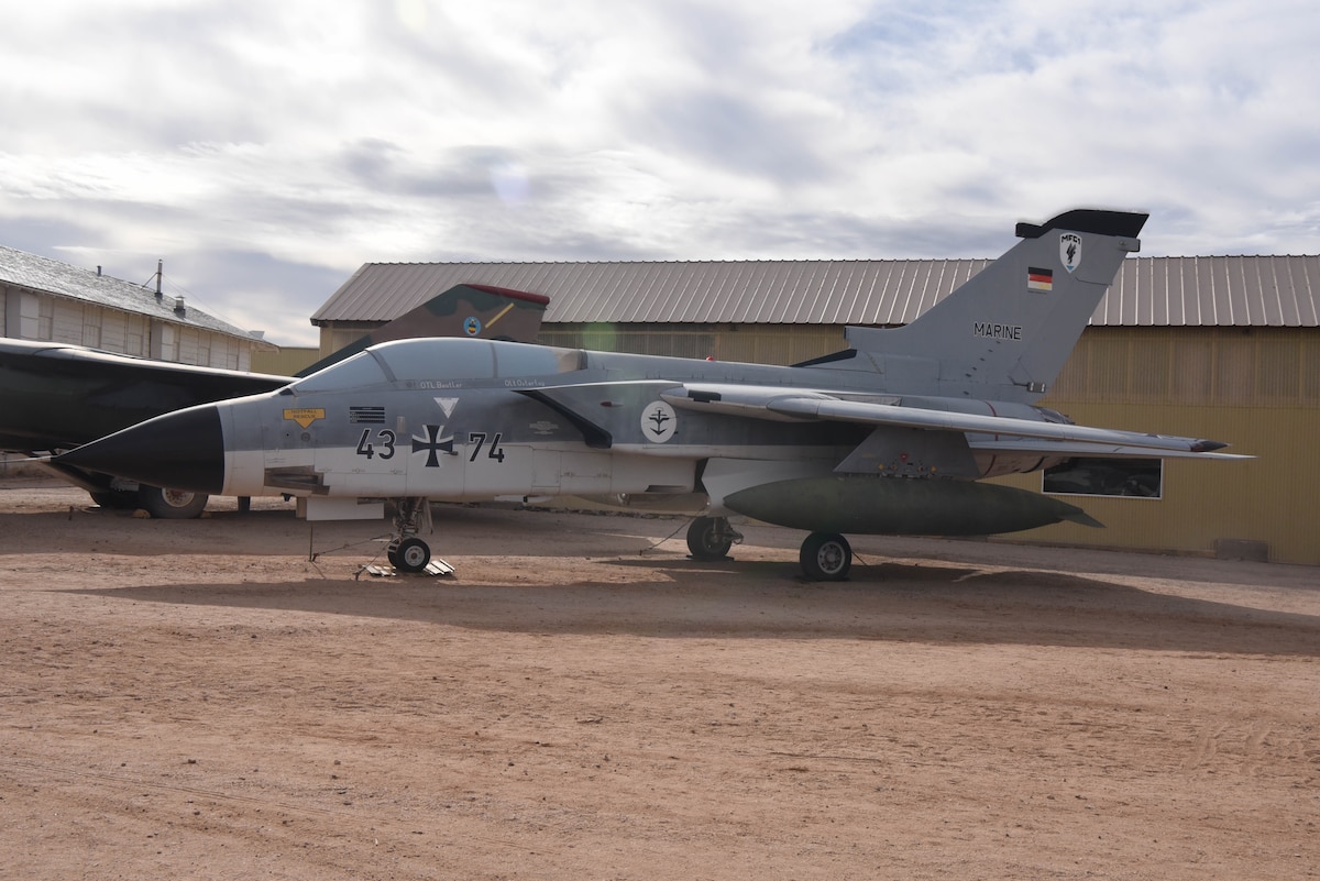 An airplane is on display in a dirt field with an aircraft to the left and building in the background.