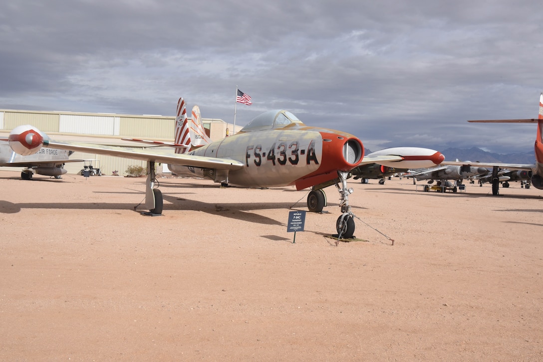 An airplane is on display in a dirt field with aircraft around it and the U.S. flag in the background.
