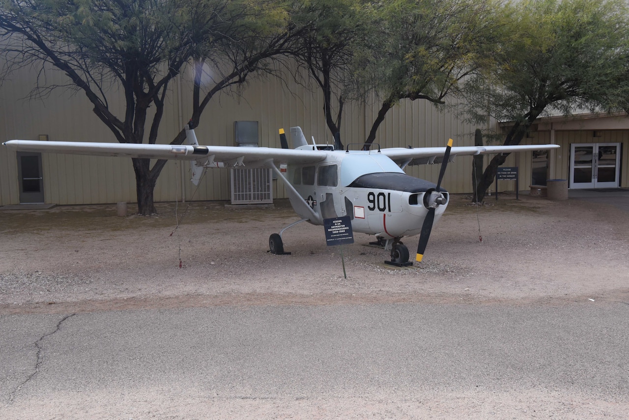 An airplane is on display in a dirt field with trees and a large building in the background.