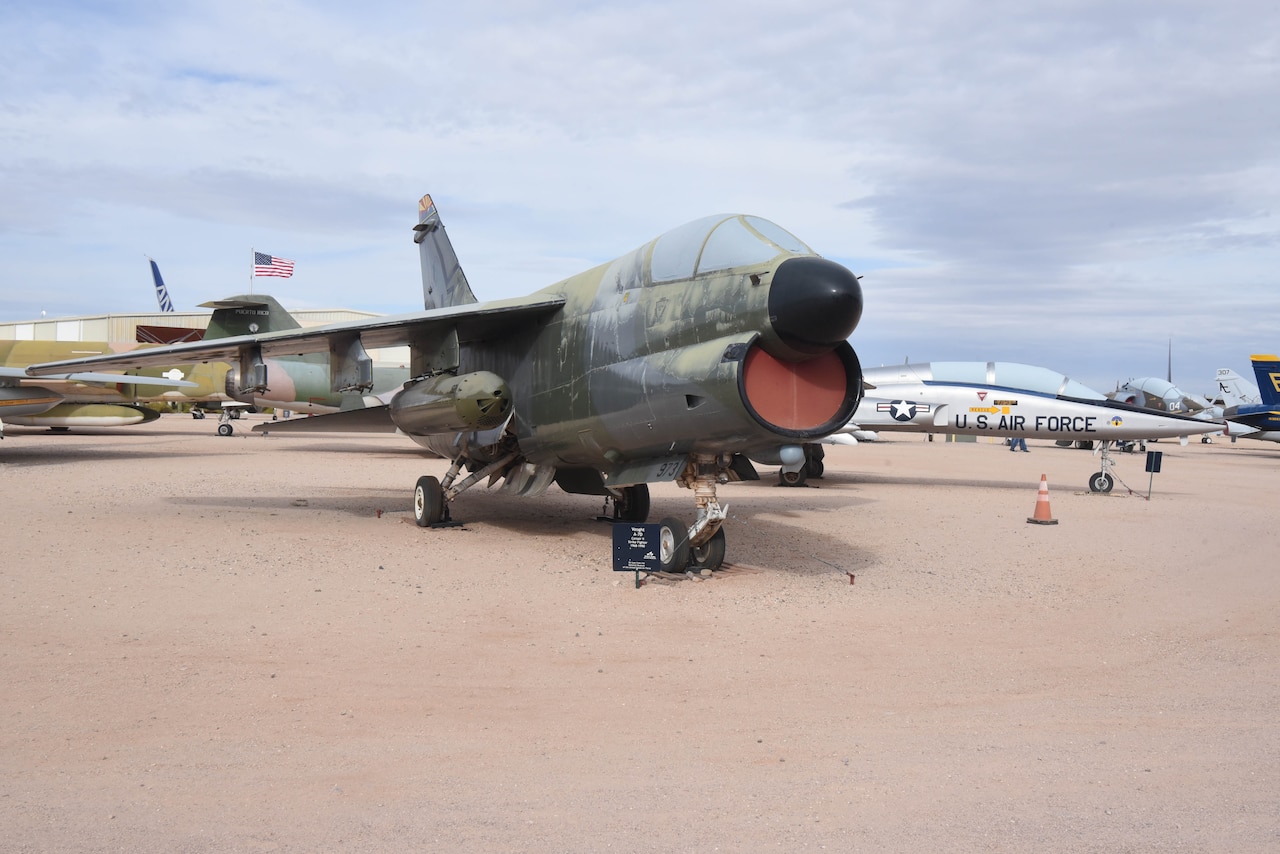 An airplane is on display in a dirt field with aircraft around it and a U.S. flag in the background.
