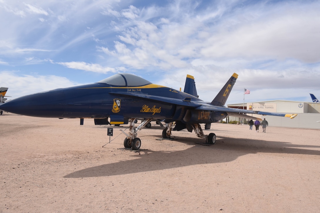 A jet is on display in a dirt field with people walking toward a large building in the background.