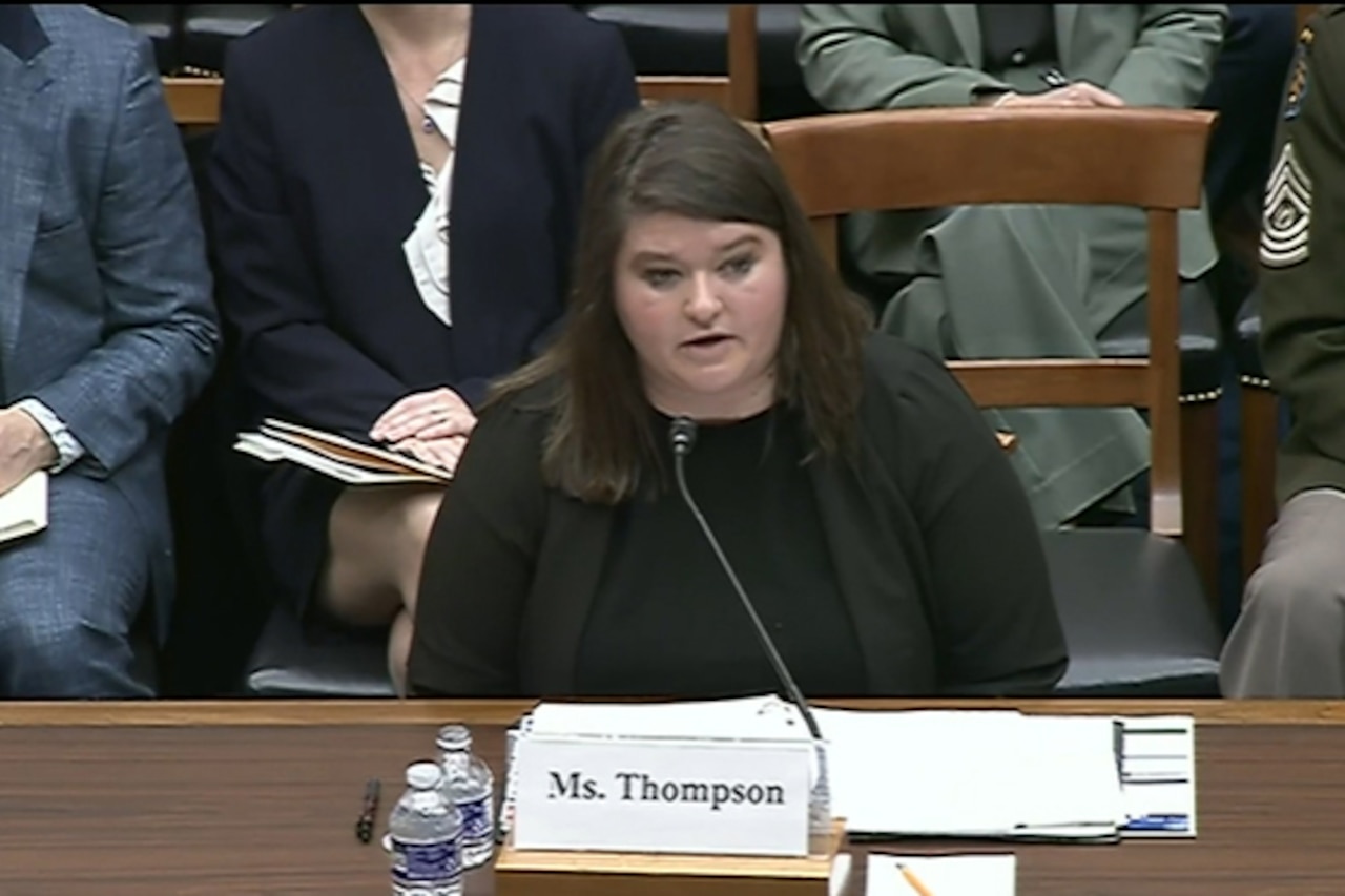A woman in business attire speaks into a microphone while seated behind a desk.