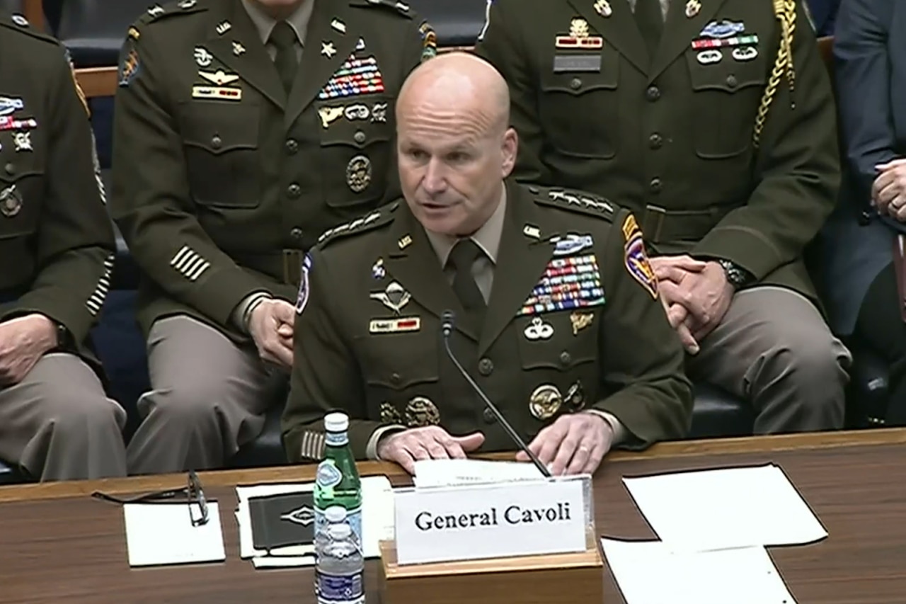 A man in a military uniform speaks into a microphone while seated behind a desk. Three soldiers in military uniforms and one person in business attire sit behind him.