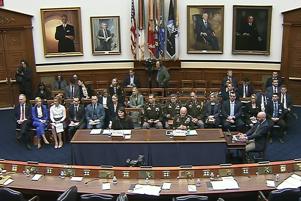 A group of people in business attire and military uniforms sits in a room with flags in the background and portraits hanging on the walls.