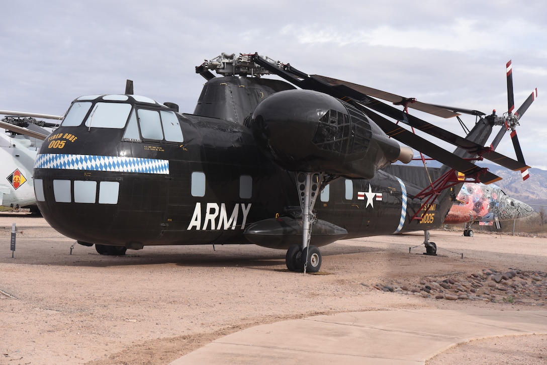 A large helicopter sits in a dirt field with a building on the right and two aircraft behind it on the left.