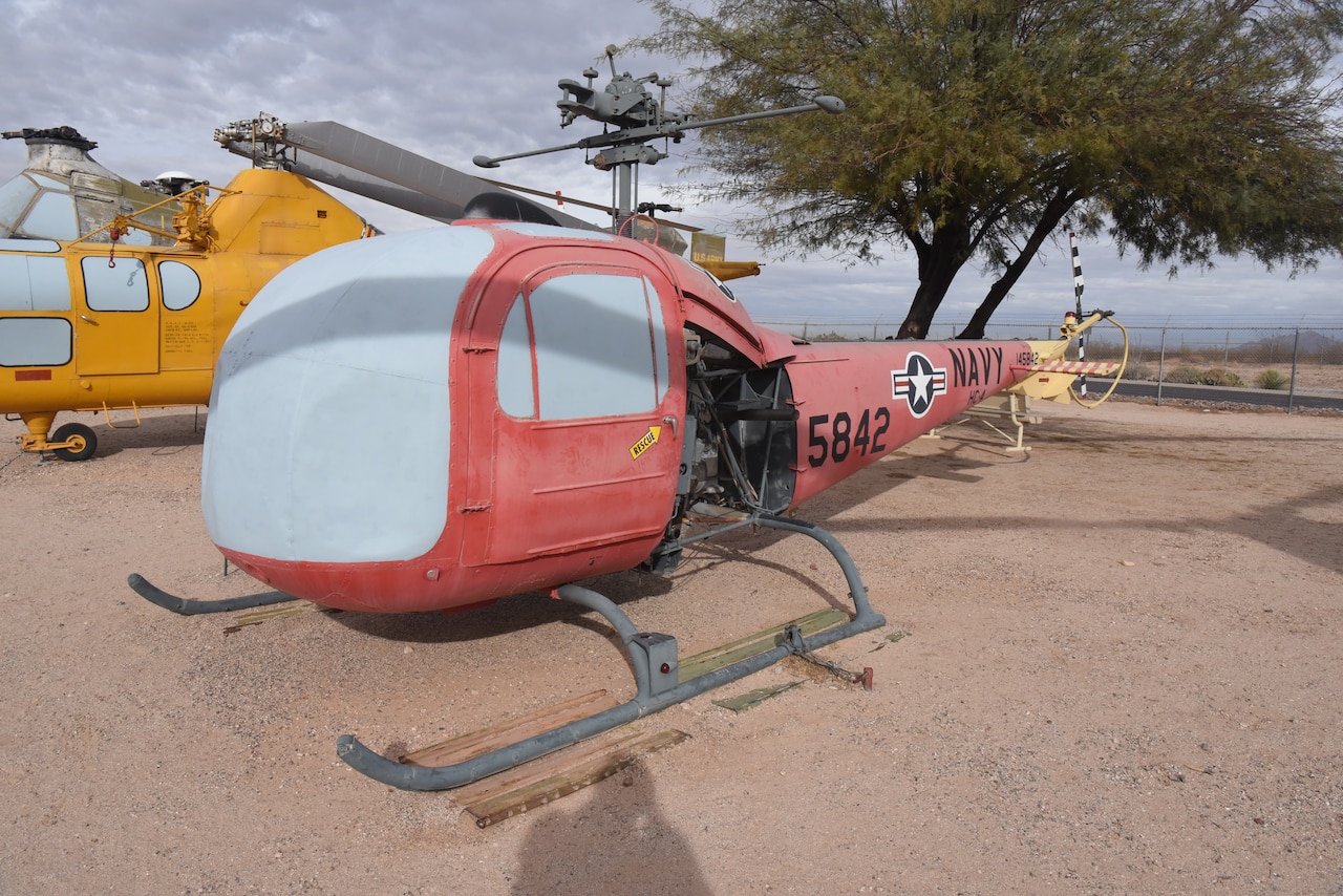 A helicopter is on display in a dirt field with an aircraft on the left and a tree in the background.