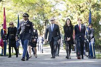 Second Gentleman of the United States Doug Emhoff and family are walking toward the camera and being escorted by an Army officer who is dressed in a dark blue service uniform. There are service members holding flags in a cordon on the sides of the walkway.
