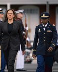 U.S. Vice President Kamala Harris is walking toward the camera next to an Army officer who is dressed in a dark blue service uniform.